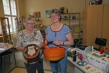 Zwei Frauen auf einem Kirchenbasar, halten einen Korb und einen Pinguin-Hocker.